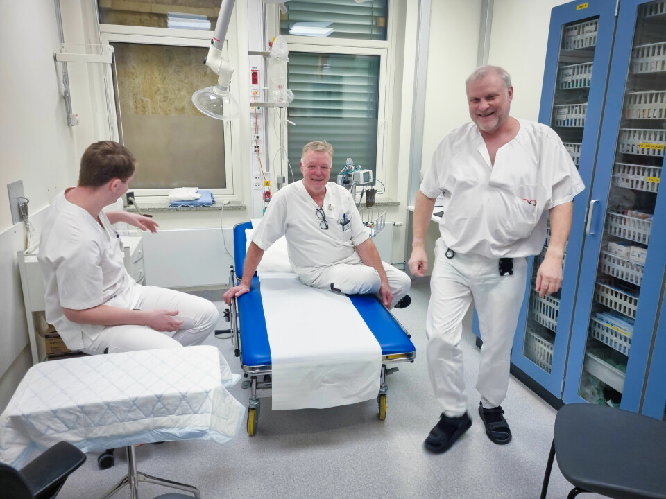Tre menn i sykepleieruniform prater og ler i et undersøkelsesrom.