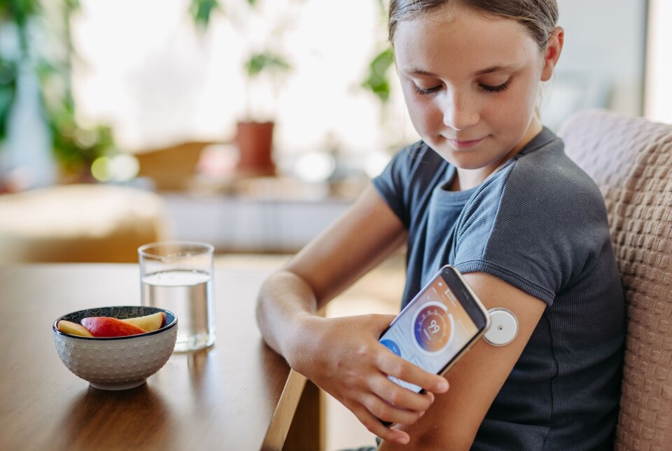 A total of around 3,500 Norwegian children and adolescents have diabetes in Norway. A new study shows that they have achieved more stable blood sugar levels, largely thanks to new technology. Jente som følger med på sitt eget blodsukker på en mobilapp. Hun sitter ved bordet. Hun har et glass vann og en skål med frukt foran seg.