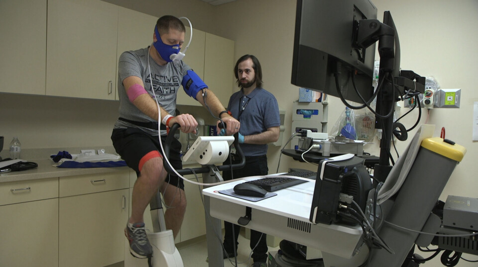 A man with ME/CFS takes a CPET test in connection with research on what happens to ME patients during physical exertion. The experiment was conducted in 2019 at the National Institute of Neurologic Disorders and Stroke at the National Institutes of Health in the USA. En mann i shorts og t-skjorte sykler på en treningssykkel i et laboratorium. Han har måleutstyr festet til armene og over nesen og munnen. En forsker står ved siden av.
