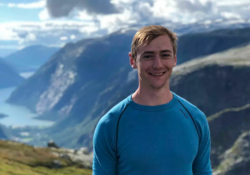 A young man with short light brown hair is smiling, dressed in a blue long sleeved top. He is standing outside, with mountains and a fjord in the background.
