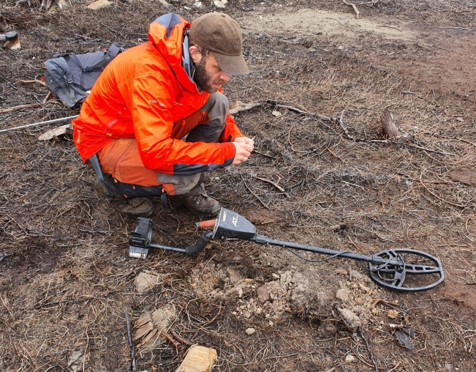 Harald Bugge Midthjell at one of the fire pits in Nesjøen, central Norway. Harald Bugge Midthjell ved et arkeologisk funn av et ildsted i Nesjøen. Han sitter på huk og er ikledd oransje turklær med en metalldetektor ved siden av.