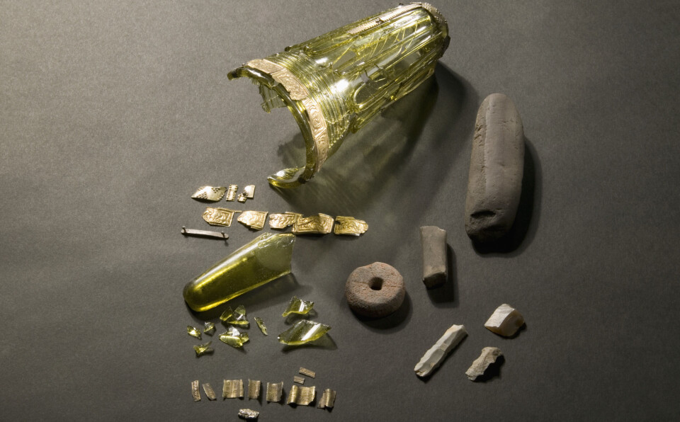 Bøckman found another glass during the excavations, but only the bottom remained. It's the glass piece that looks like a tube. According to Bøckman, this glass was likely shattered and transported away during the processing of the soil. Gulgrønt, avlangt beger med gullbånd øverst. Begeret er delvis knust, og flere biter ligger ved siden av.