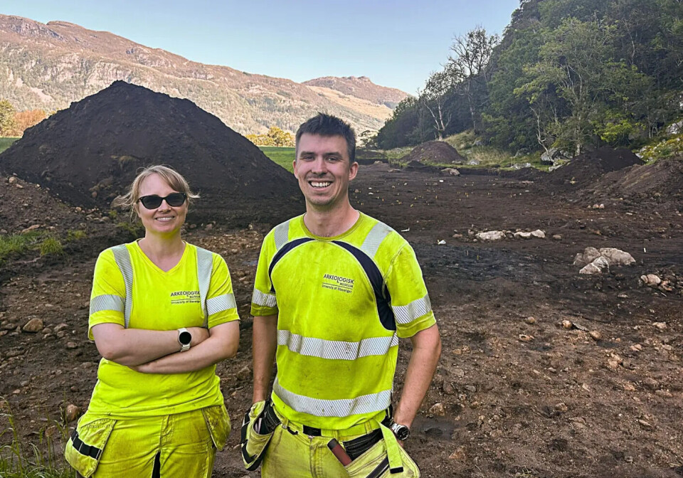 Field archaeologists Mari Krogstad Samuelsen and Ola Tengesdal Lygre first discovered the Viking Age treasure. A man and a woman pose in front of a mound of soil in yellow work clothes.
