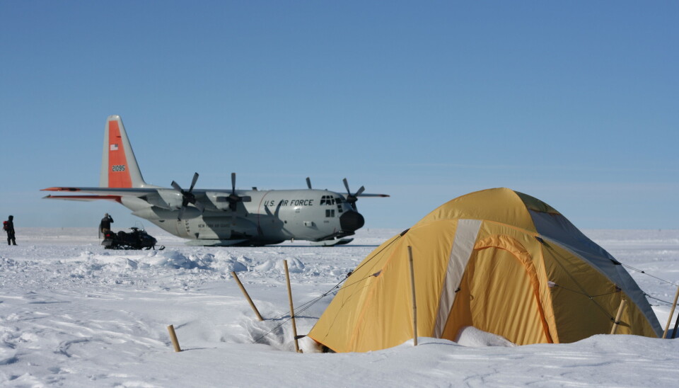 The researchers live in tents, and a plane comes once a month. Et lasteskip i bakgrunn, med et telt foran. Alt er dekt i snø.