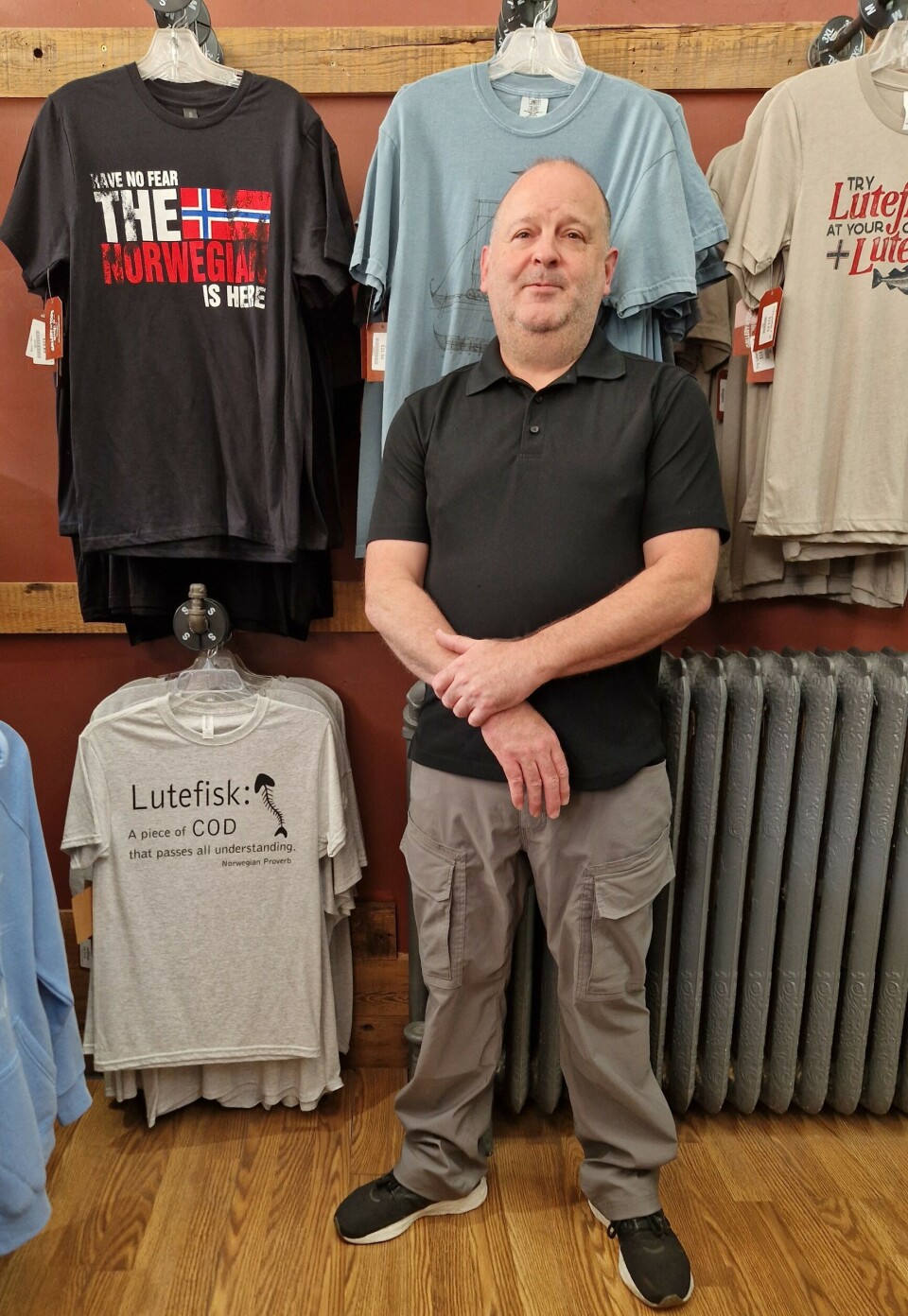 Mike Cline runs a T-shirt shop in Decorah, Iowa. Many of them have slogans about Norwegians and lutefisk. 'They sell well. And not just to Norwegian-Americans. The whole area is Norwegian, so many people are married into Norwegian families. They think the T-shirts are funny,' says Cline. Bilde av Mike Cline foran t-skjorte med påskrift: Frykt ikke. Nordmennene er her. Og t-skjorter med tekst om lutefisk.