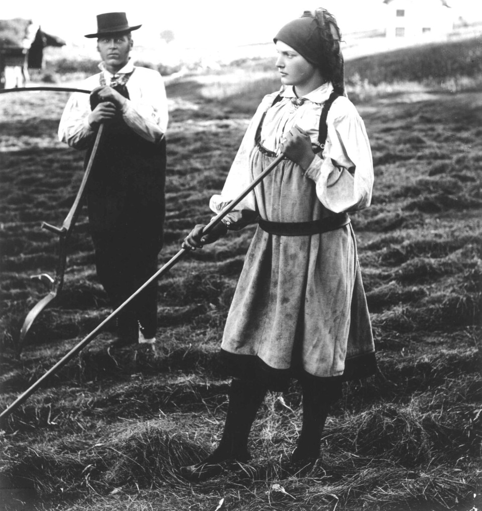 Folk costumes served as both festive clothing and everyday wear. The newest and least worn garments were used for Sunday church. At home, people changed into the next-best items. As garments wore down, they dropped in status, eventually ending as work clothes, according to Bjørn Sverre Hol Haugen. This photo was taken in Bygland in Setesdal in 1888. Mann og kvinne i folkedrakt med ljå og rive på jorde.