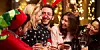 Three women and two men in Christmas jumpers and headgear laugh while drinking.