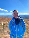 Forsker Maja Eilertsen avbildet på fjelltur. Hun har knallblå jakke, og står i terreng med høstfarger og flott utsikt.