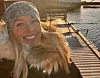 Woman in winter coat smiles on a sunlit wooden pier by the water.