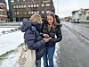 Two teens in puffer jackets look at their phones on a snowy pavement in a city.