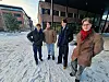Four male students in winter coats standing outside on the snow between brick buildings.