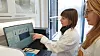 Two female researchers in lab coats examine graphs on a computer screen.