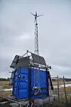 Blue container with rooftop solar panels and a wind turbine in an open field