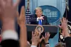 Donald Trump standing at a podium as reporters raise their hands during a press briefing