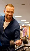 Man in a blue top curls a dumbbell in a gym with equipment behind him