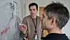 Two colleagues stand at a glass whiteboard covered with diagrams and red annotations.
