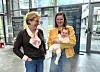 Two women standing indoors, one in a yellow jacket holds a baby.