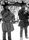 Old black-and-white photo of two men in traditional winter dress stand on snow in a rural, mountainous setting.