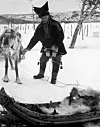 Old black-and-white photo of a man with a reindeer and sled in a snowy landscape.