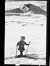 Old black-and-white photo of a small child on skis holding poles in a snowy field below a high mountain