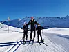 Three cross-country skiers stand on a groomed snowy track in front of mountains.