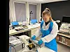 Woman in a white lab coat and blue apron and gloves handles equipment at a lab workstation.