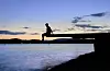 Silhouetted person sitting on a jetty above the water at dusk with calm water and distant lights.