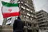 Person holding an Iranian flag facing a building damaged by bombs.