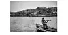 Fisherman standing in a wooden boat on calm water near a rocky shore