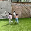 Three children play hide and seek on artificial grass beside a wooden wall.