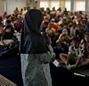 Girl in headscarf speaks into a microphone before children seated in a large room