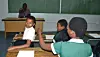 Students at wooden desks raise their hands toward a teacher standing in front of a green chalkboard.