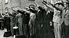 Men lined up on a street give raised-arm salutes beside a brick building.