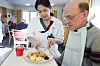 Care worker cuts food on a plate while assisting an older man wearing a bib at a dining table.