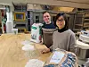 Two people in a bakery kitchen stand by flour samples and large flour bags on a wooden bench