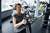 Older woman using a suspension trainer in a gym with cardio equipment behind her.