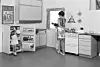 Black-and-white photo of a woman cooking in a 1950s kitchen while a young girl stands by an open fridge.