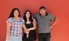 Three smiling young people pose together in front of a bright red wall.