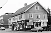 Black and white view of Knasken frukt og tobakk on a corner street with parked cars and adjacent buildings.