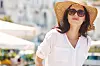 Young woman in straw hat and sunglasses walking past outdoor cafés in a sunny city.