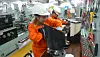 Two engineers in orange coveralls work on machinery in an engine room workshop.