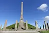 Tall stone obelisk on a grass-covered mound with flanking standing stones under a blue sky.