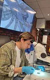 Scientist in gloves using a microscope with the specimen magnified on a screen behind