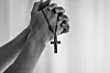 Black-and-white close-up of folded hands holding a rosary cross in prayer.