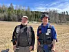 Two workers in safety gear stand in a harvested field before a tree line