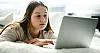 Young woman on a bed looking at a laptop screen in a bright home setting.