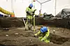 Archaeologists in high-vis gear excavate a trench inside a tented dig site.