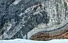 Geologists on a glacier below a steep cliff with dark rock bands and pale marble layers.