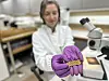 Gloved hands holding a small gold ornate item next to a microscope in a lab.