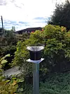 Netatmo rain gauge mounted on a post in a garden with shrubs, a house and cloudy sky behind.